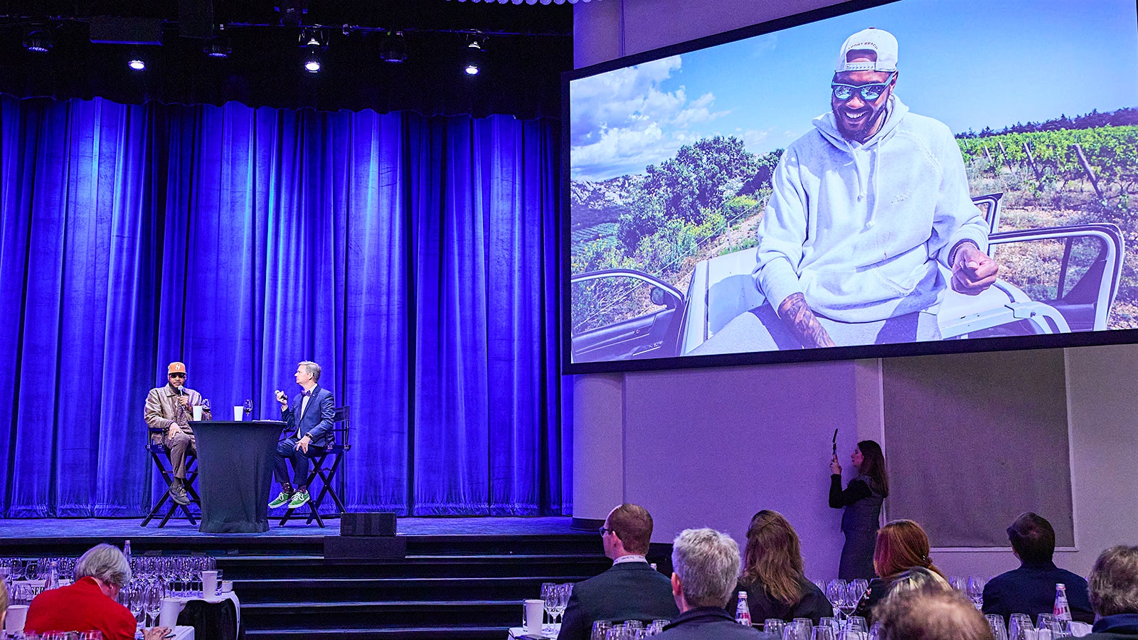 Carmelo Anthony, onstage with Wine Spectator senior editor Mitch Frank, and pictured on screen sitting on the roof of a truck in a vineyard.